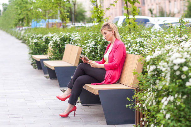 Side view of young successful serious businesswoman in elegance style holding tablet pc and reading online with focused serious face while sitting on bench in park - Fotoğraf, Görsel