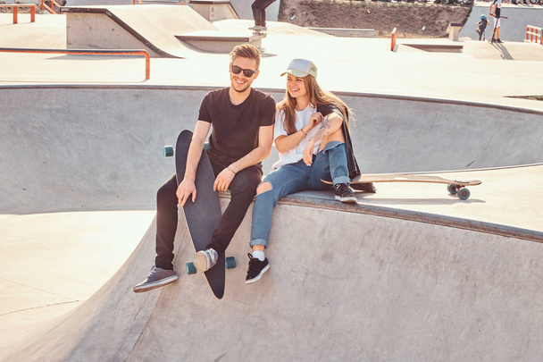 Sorrindo casal feliz de estudantes estão sentados no parque de skate com seus longboards
 - Foto, Imagem
