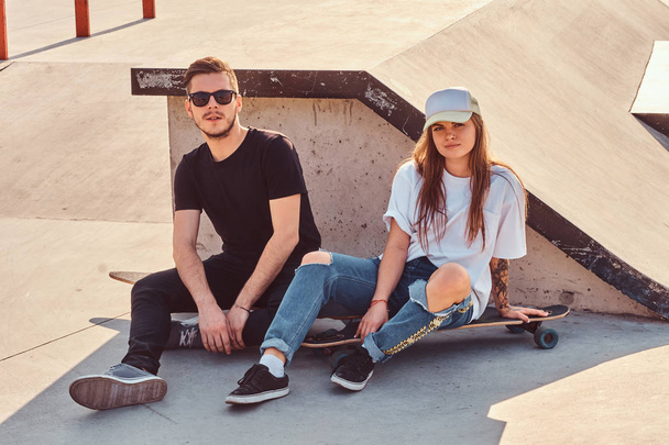 若いトレンディーなカップルは彼らの長いボードと日当たりの良いスケートパークに座っています - 写真・画像