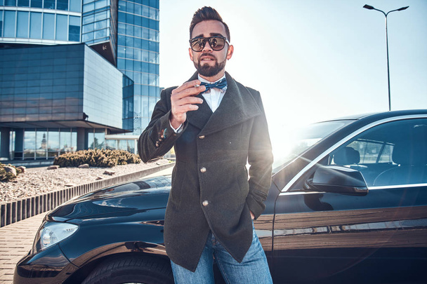 schöner Mann im Mantel steht neben seinem neuen Auto, während er auf dem Parkplatz raucht. - Foto, Bild