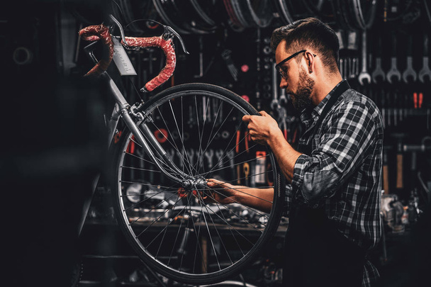 Привлекательный задумчивый мужчина в защитных очках ремонтирует велосипед в собственной мастерской
 - Фото, изображение