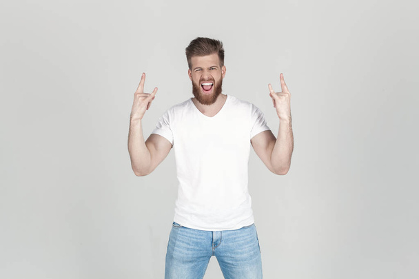 Hombre exitoso con barba de jengibre exuberante. gritando con las manos levantadas con una señal de roca, de pie delante del fondo blanco
 - Foto, Imagen