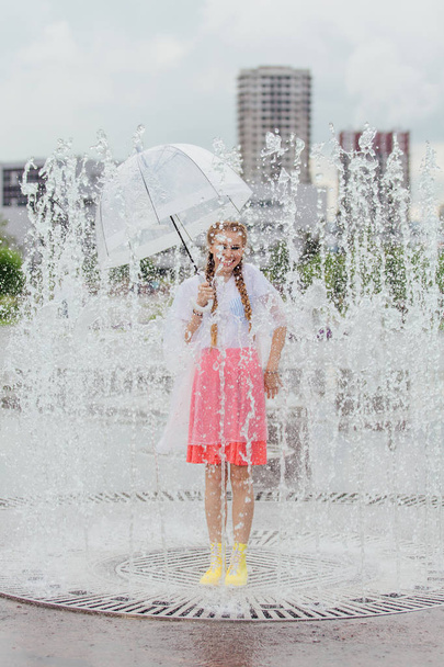 Young wet pretty girl with two braids in yellow boots and with transparent umbrella stands inside of fountain. - Photo, Image