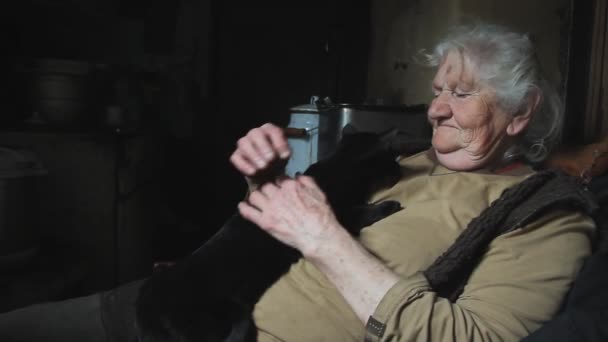 Een oude vrouw met grijs haar houdt haar zwarte kat in haar armen, speelt met haar, glimlacht, woont in een verlaten dorp, selectieve focus - Video