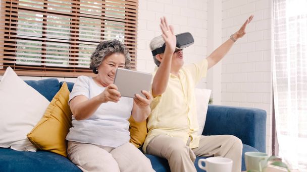Azjatycka para starców korzystająca z tabletu i symulatora rzeczywistości wirtualnej grająca w gry w salonie, para czująca się szczęśliwa korzystając z czasu spędzonego razem na kanapie w domu. Styl życia Starsza rodzina w domu koncepcja. - Zdjęcie, obraz