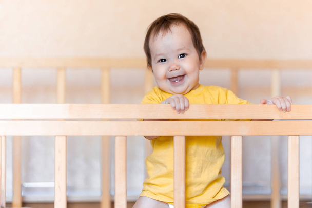 fröhliches kleines Mädchen mit dunklen Haaren, lachend und lächelnd in gelbem T-Shirt, gemischt aus Kasachen und Deutschen, die im Kinderbett stehen - Foto, Bild