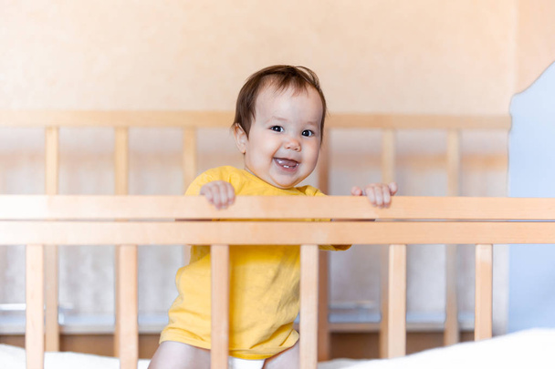 Gelukkig baby meisje met donker haar lachen en glimlachend in geel t-shirt gemengde race Kazachse en Duitse staande in Cot wieg - Foto, afbeelding
