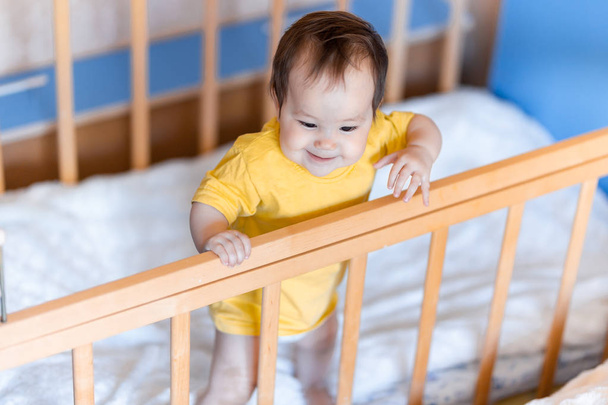 Happy baby girl with dark hair laughing and smiling in yellow t-shirt mixed race kazakh and german standing in cot crib - Photo, Image
