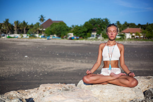 young beautiful woman in white bikini practicing yoga sitting on stone sea coast with tropical view in background. she sits in the lotus position. her palms are folded for prayer. Meditation - Fotoğraf, Görsel
