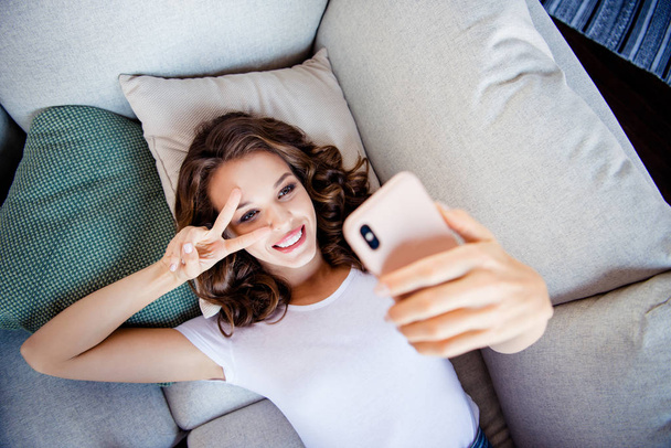 Top above high angle view portrait of her she nice-looking attractive lovely lovable shine sweet cheerful cheery wavy-haired girl lying on divan taking selfie showing v-sign indoors - Foto, Imagem