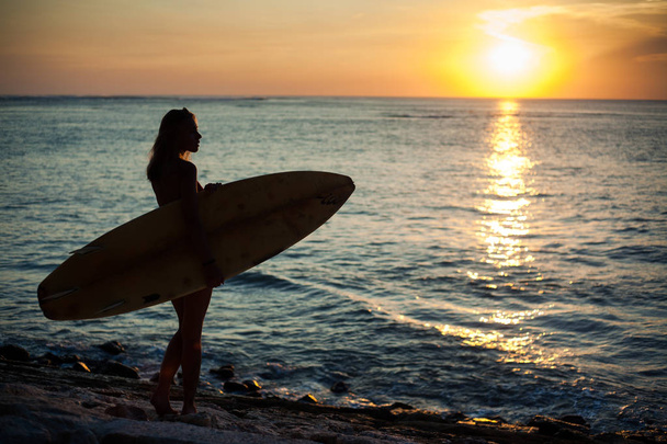 silhouette of a girl in backlight holding a surfboard. Sunset on the sea coast. sun touchdown - Fotoğraf, Görsel