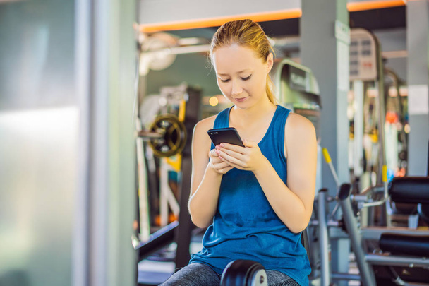 Jeune femme utilisant le téléphone pendant l'entraînement à la salle de gym. Femme assise sur la machine d'exercice tenant le téléphone portable
 - Photo, image