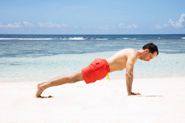 Shirtless Young Man In Red Shorts Exercising On Sandy Beach In Front Of Sea - Foto, Bild