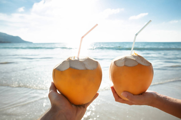 Gros plan de la main du couple tenant la noix de coco avec de la paille à boire devant la mer idyllique
 - Photo, image