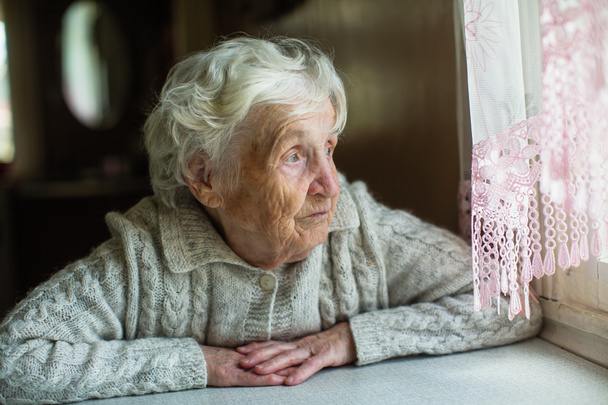 Μια ηλικιωμένη γυναίκα φαίνεται δυστυχώς έξω από το παράθυρο. Φροντίδα για τους μοναχικούς συνταξιούχους. - Φωτογραφία, εικόνα