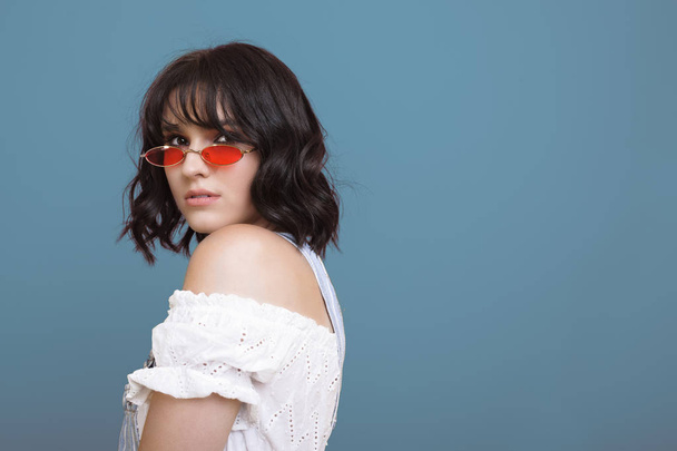 Close up portrait of a beautiful caucasian woman looking seriously over her shoulder wearing fashionable red sunglasses isolated over blue background. - Fotó, kép