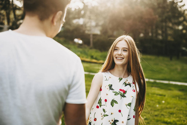 Portrait of lovely caucasian woman with long red hair and freckles looking at his boyfriend laughing against sunset light while holding hands while dating. - Foto, immagini