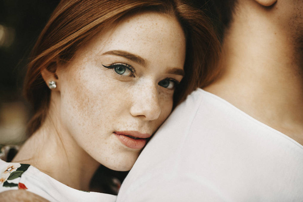 Close up portrait of a amazing red haired woman with freckles and green eyes looking at camera while leaning her head on her boyfriend back outside while dating. - Photo, Image