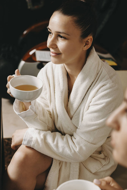 Portrait of a beautiful young woman drinking tea dressed in a bathrobe in a wellness spa center after massage. - Photo, Image