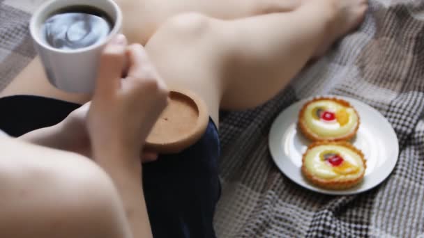 女の子はベッドで朝のコーヒーとケーキで目を覚ます。居心地の良い青いローブでベッドでコーヒーを飲む美しい若い女性. - 映像、動画
