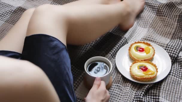 Το κορίτσι ξυπνάει με πρωινό καφέ και τούρτα στο κρεβάτι. Όμορφη νεαρή γυναίκα πίνοντας καφέ στο σπίτι στο κρεβάτι της με μια ζεστή μπλε ρόμπα. - Πλάνα, βίντεο