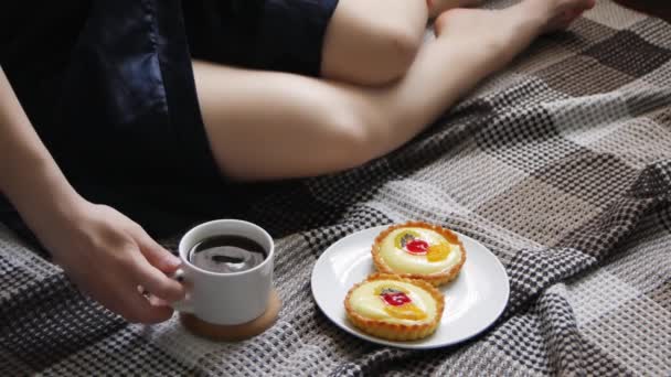 Το κορίτσι ξυπνάει με πρωινό καφέ και τούρτα στο κρεβάτι. Όμορφη νεαρή γυναίκα πίνοντας καφέ στο σπίτι στο κρεβάτι της με μια ζεστή μπλε ρόμπα. - Πλάνα, βίντεο