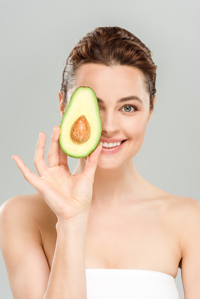 happy woman holding half of organic avocado while covering eye isolated on grey  - Foto, Bild