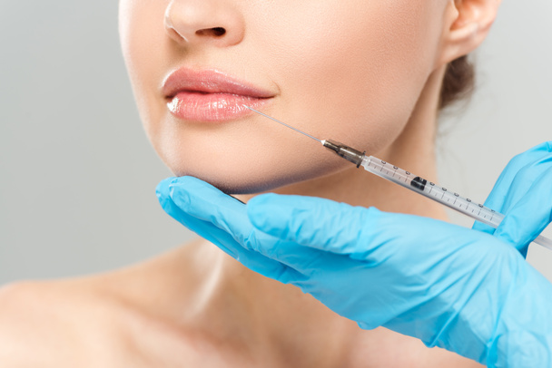 cropped view of cosmetologist in blue latex gloves holding syringe near face of woman isolated on grey  - Фото, изображение