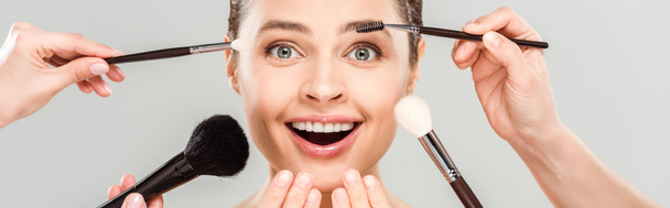 tiro panorâmico de maquiadores segurando escovas cosméticas perto da mulher excitada isolada no cinza
  - Foto, Imagem
