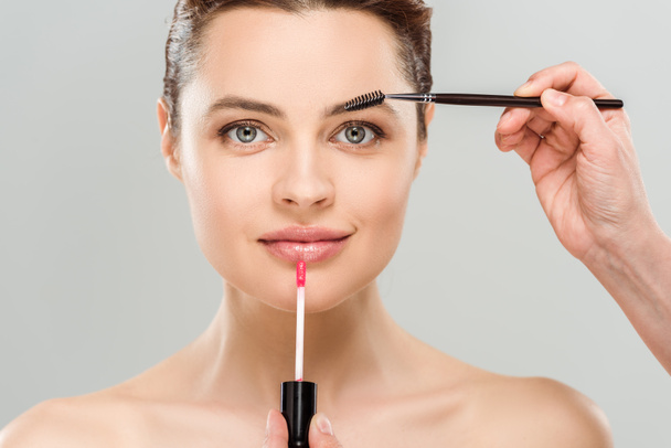 cropped view of makeup artist holding mascara brush and lip gloss near naked woman isolated on grey  - Foto, Bild