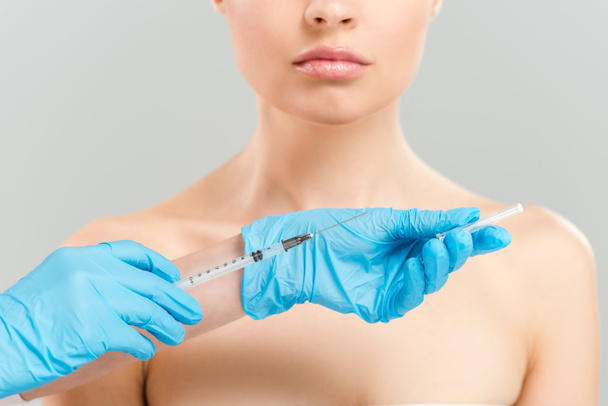 обрезанный вид косметолога в голубых латексных перчатках, держащего шприц рядом с обнаженной женщиной, изолированной на серой
  - Фото, изображение