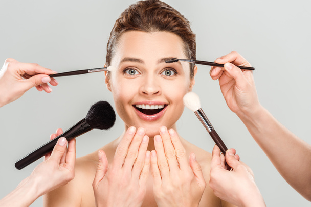 cropped view of makeup artists holding cosmetic brushes near naked excited woman isolated on grey  - Fotografie, Obrázek