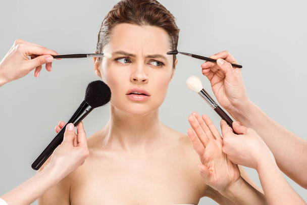 vista cortada de maquiadores segurando escovas cosméticas perto de mulher descontente nu isolado em cinza
  - Foto, Imagem