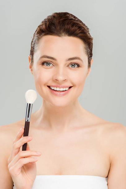 mujer alegre sosteniendo cepillo cosmético y sonriendo aislado en gris
  - Foto, Imagen
