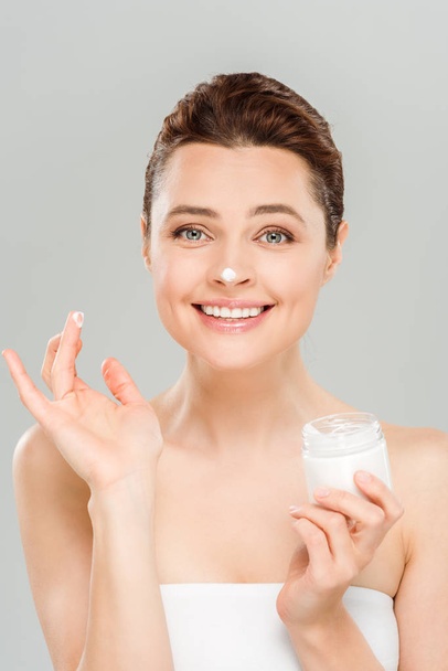 cheerful woman applying face cream and smiling isolated on grey  - Photo, Image