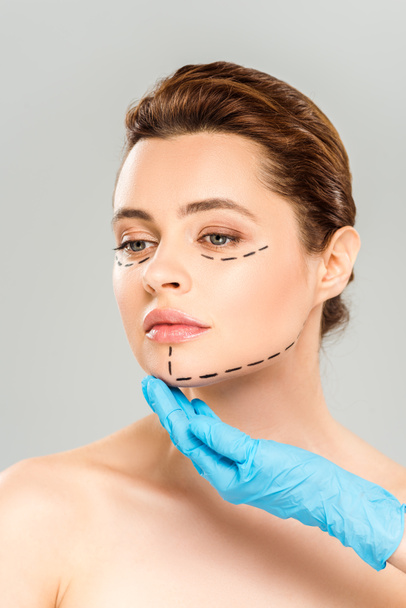 灰色で隔離されたマークされた顔を持つ女性の顔に触れる形成外科医のトリミングされたビュー  - 写真・画像