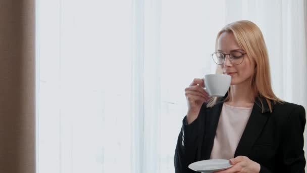 Young attractive middle-aged blonde woman in a black business suit and glasses in a hotel room. Stands by the window, drinks coffee from a white cup, looks out of the window and smiles. - Video