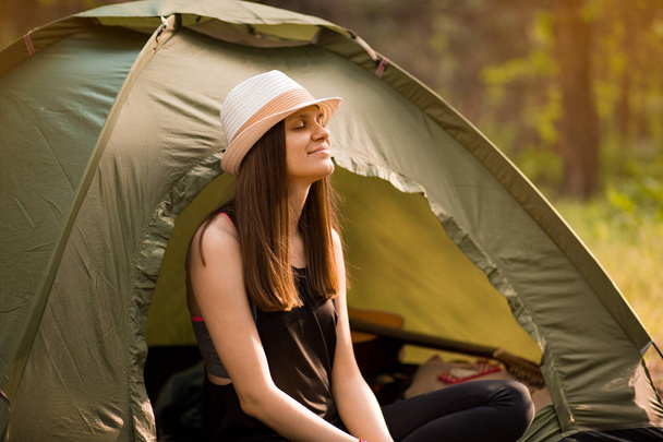 Young slim hiker woman sitting at small tourist tent enjoying beautiful nature forest on bright summer morning. - Photo, image