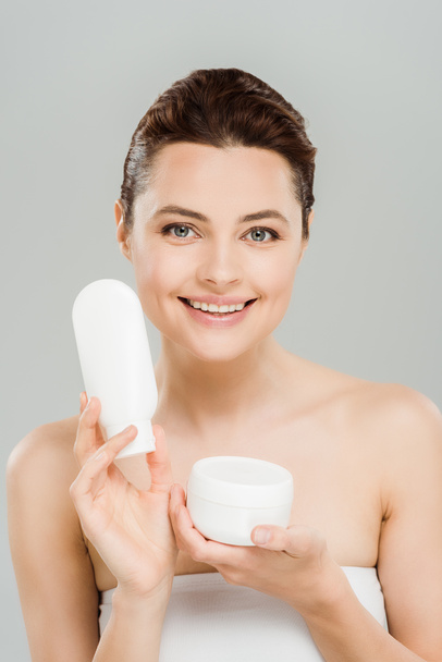 cheerful woman holding container with cosmetic cream and bottle of body lotion isolated on grey  - Photo, Image