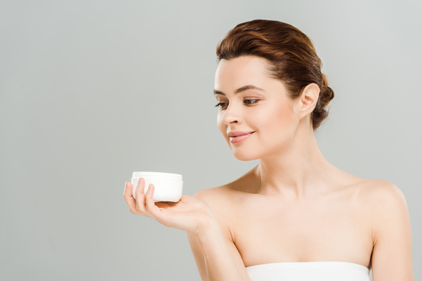 cheerful woman looking at container with cosmetic cream isolated on grey  - Photo, Image