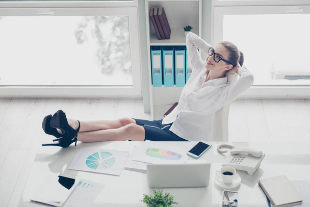 Close up photo beautiful she her business lady hands arms behind head joy fun overjoyed vacation thoughts telephone notebook papers legs heels table wear specs formal-wear shirt skirt bright office - 写真・画像