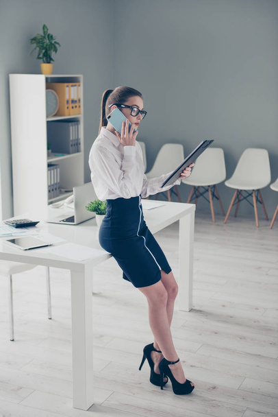 Vertical full length side profile body size photo beautiful she her business lady hands arms reader text report speak telephone lean table wear specs high-heels formal-wear shirt skirt bright office - Foto, imagen