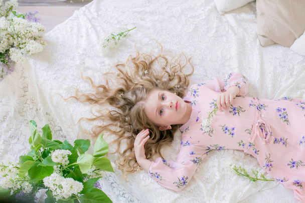 Cute little girl with blond hair in a beautiful dress in a spring studio with lilac flowers - Photo, Image