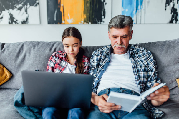 Grandfather reading book while granddaughter using laptop - Φωτογραφία, εικόνα
