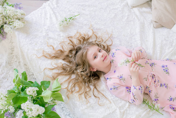 niedliches kleines Mädchen mit blonden Haaren in einem wunderschönen Kleid in einem Frühlingsstudio mit lila Blumen - Foto, Bild
