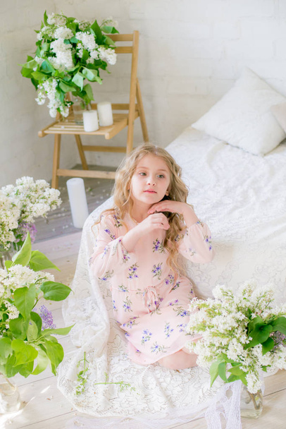 Cute little girl with blond hair in a beautiful dress in a spring studio with lilac flowers - Foto, immagini