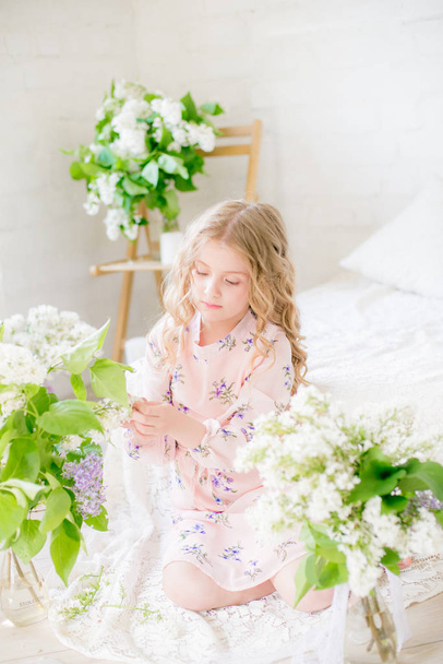 Cute little girl with blond hair in a beautiful dress in a spring studio with lilac flowers - Foto, imagen