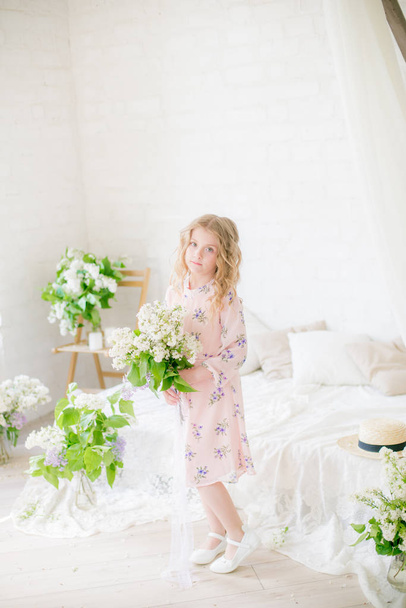 Cute little girl with blond hair in a beautiful dress in a spring studio with lilac flowers - Fotó, kép