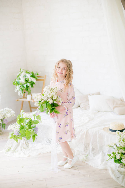 Cute little girl with blond hair in a beautiful dress in a spring studio with lilac flowers - Fotografie, Obrázek