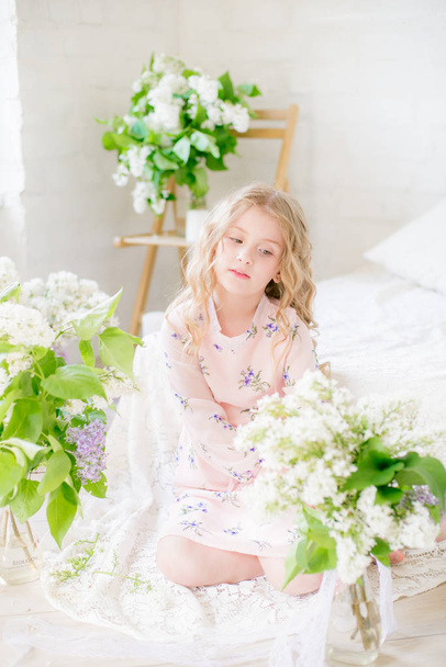Cute little girl with blond hair in a beautiful dress in a spring studio with lilac flowers - Фото, изображение